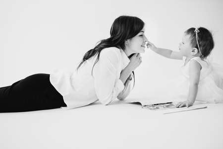 Black and white photo of happy mom laying on the floor with her baby daughter and reading the magazine, little girl gently touching mother nose, maternity concept. High quality photoの写真素材