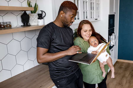 Smiling multiethnic parents relax at home with cute little biracial son using laptop together, happy international family with small boy child stay in kitchen watch video or talk with family onlineの写真素材