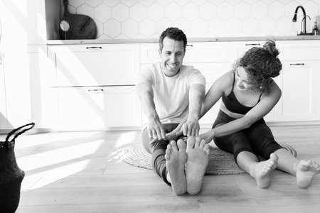 Black and white portrait of barefoot happy couple sitting warming up in sportswear on the floor at home before work. Smiling man and woman stretching isolated on the carpet together.High quality photoの写真素材