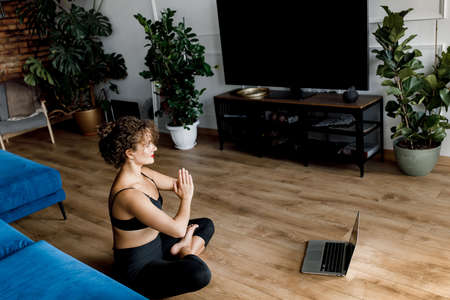 Back view of millennial woman doing yoga exercise and watching tutorial lesson on laptop computer at home. Beautiful female sitting on the floor near sofa listening meditation isolated.の写真素材