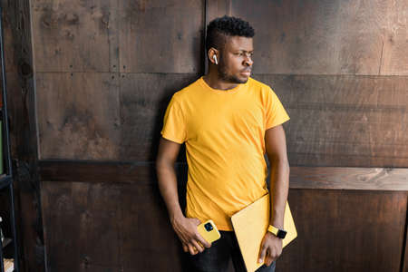 Serious focused afro american freelancer is standing near the wall, with yellow laptop, smart phone...