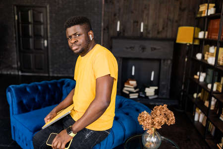 Serious millennial african american business man entrepreneur standing at home at stylish sofa, confidently looking at camera, hold yellow laptop and smartphone after working on project or studyingの写真素材