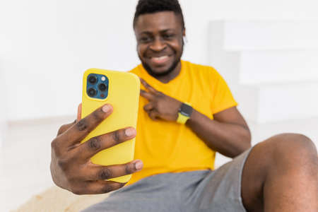Joyful young african american ethnicity male smiles makes victory peace fingers gesture, taking selfie photo on yellow smart phone front camera isolated on white background, smartphone in focusの写真素材