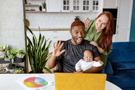Head shot happy african american mixed race family looking at yellow laptop, holding video call with relatives, friends waving in modern apartment. Joyful married couple showing baby son onlineの写真素材