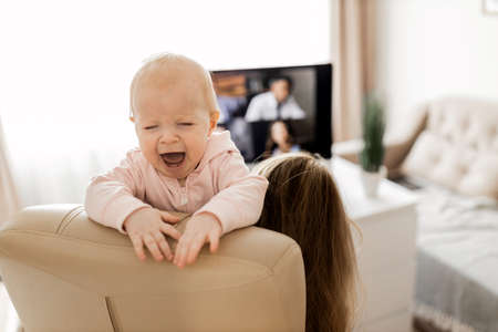 Young woman working at desk at home with computer holding crying tired infant baby holding business meeting at home, online pediatric consultation, chatting with grandparents, mothers forum conceptの写真素材