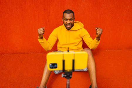 Strong Afro American sportsman is sitting in front of the smartphone camera, making video call about online sport exercises. Overjoyed winner in yellow sportswear doing victory gesture isolatedの写真素材