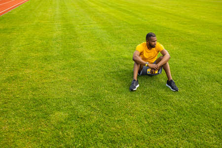 Young pensive Afro American sportive male sitting with smartphone on grass in the middle of stadium, relaxing after workout training, football soccer match. Copy space. Healthy sport lifestyle conceptの写真素材
