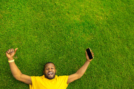 Top view of cheerful overjoyed African American male celebrating victory, successful race, game or football match. Young afro laying on the grass field stadium, holding mobile phone and show thumb upの写真素材