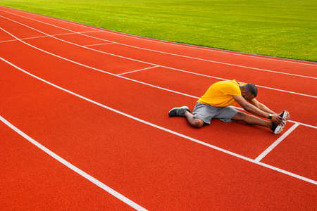 African american young male work out hard, sitting at stadium stretching legs on bright background. Professional young black man in yellow sportswear warming up body before doing exercises, training.の写真素材