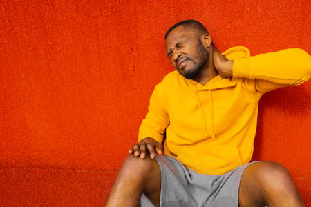 Exhausted Afro American sportsman in yellow sportswear resting after intensive self training outdoors, male runner athlete afro american race taking a break after evening exercises, having neckacheの写真素材