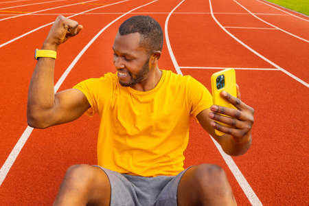 Handsome sporty Afro American millennial male in casual yellow t-shirt smiling happily and raising clenched fist, tensing muscles, feeling strong and full of energy taking photo on mobile phoneの写真素材