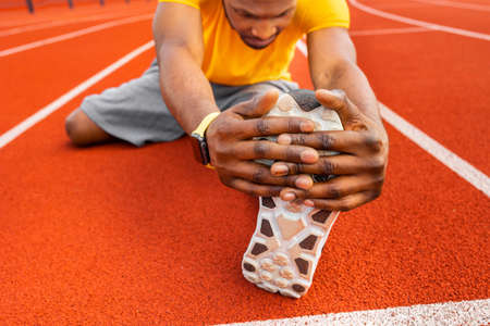 Close up african american young male sitting at stadium stretching legs on bright background....