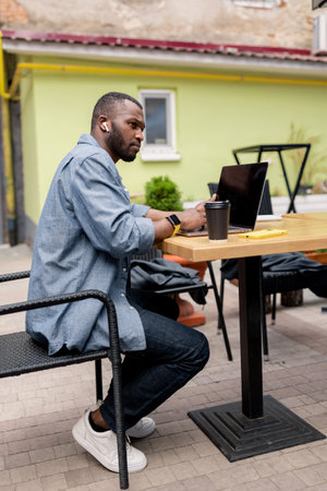 Side view of focused pensive Afro American freelancer wear wireless headphones, holding online meeting, watching educational lessons on laptop remotely online work or study in cafe, looking at screenの写真素材