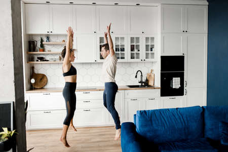 Focused sporty millennial couple having fun at home kitchen together, active excited young man and woman doing sport and jump raising hands during pandemic, looking at each other. High quality photoの写真素材