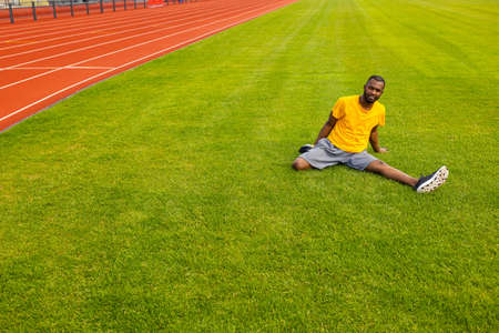 Smiling african american male in stylish sportswear sitting at the grass, trying to do a split, stretching legs. Athletic man is working out, doing warm up before training outdoors conceptの写真素材