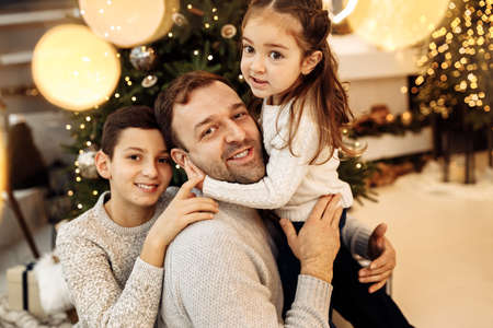 Portrait of caring father with little kids sitting near Christmas tree, smiling. Happy man with adorable daughter and joyful son celebrate New Year at home, enjoy winter holidays, happy family conceptの写真素材