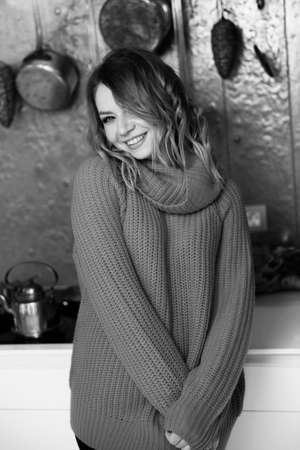 Black and white portrait of beautiful smiling woman in cozy sweater at home. Stylish young adult female standing at the kitchen, looking at the camera, preparing for dinner, winter holidays conceptの写真素材