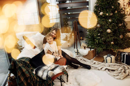 Beautiful stylish woman with little son sitting near Christmas tree at home, smiling and cuddling....