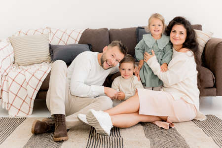 Happy beautiful parents sitting near the sofa with joyful little kids, smiling, spending weekends at home. Charming father with pretty mother playing with cute daughter and son, family conceptの写真素材