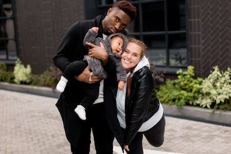 Portrait of stylish multiracial family with cute toddler at the park, smiling and hugging. Young beautiful parents with little son having a walk, looking at the camera, weekends outdoor concept.の写真素材