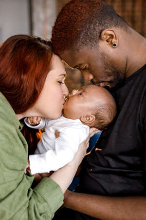 Happy multiracial mixed family gently holding newborn baby. Red haired caucasian mom looking at her...