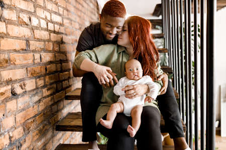 Portrait of mixed race smiling family sit on the stairs. Beautiful mother and smiling father holding baby boy. Joyful parents looking gently at their son spend weekends at home, childcare concept.の写真素材