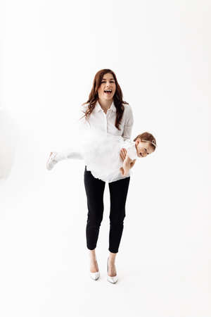 Happy family indoors in studio. Attractive young mother wearing high heels and white blouse throws the baby girl up, laughs and plays together on the white background. high quality photoの写真素材