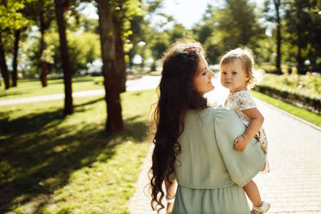 Adorable woman with little baby girl at the park, lovely daughter sitting on mothers hands, smiling, mom gentle kiss cute toddler, enjoy tender family moments, parenting concept. high quality photoの写真素材