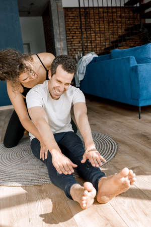 Beautiful curly-hair female trainer helps her boyfriend warm up at home during quarantine. Very...