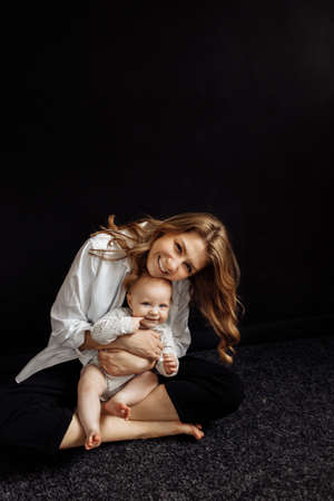 Beautiful mom is sitting in the studio and holding her little daughter, looking at the camera, smiling. Beautiful young woman and adorable baby girl posing at the black background.の写真素材