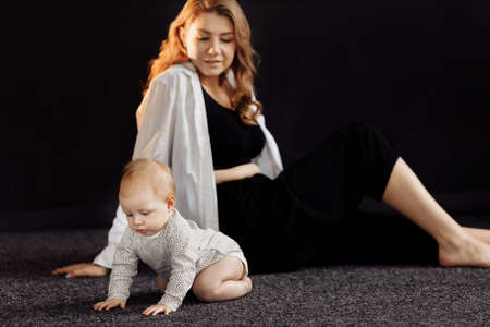 Attractive stylish woman sitting and holding adorable baby in her arms. Beautiful young mother and her lovely daughter looking confidently at the camera, posing in studio, photoshoot concept.の写真素材