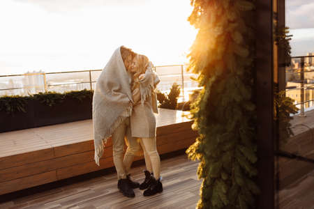 Beautiful marriage couple wrapped in blanket stand at the balcony, handsome man hug gorgeous woman at the sunrise, enjoy tender moments, winter holidays, Christmas morning concept. high quality photoの写真素材