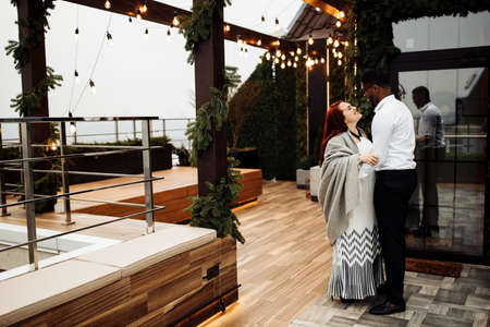 Happy couple stand at the balcony, handsome African American man hug gorgeous white woman wrapped in...