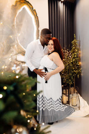 Wonderful couple stand near Christmas tree at home, caring husband hug pregnant wife, young family enjoy happiness, spend winter holidays together, New Year celebration concept. high quality photoの写真素材