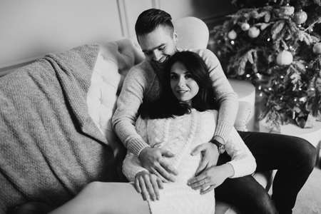 Happy couple sit on the sofa near decorated Christmas tree, beautiful pregnant wife with caring husband cuddling, spend winter holidays together, New Year celebration concept. high quality photoの写真素材