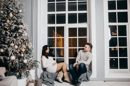 Beautiful couple sit near decorated Christmas tree, cuddling, beautiful wife with loving husband spend winter holidays together, enjoy tenderness, New Year celebration concept. high quality photoの写真素材