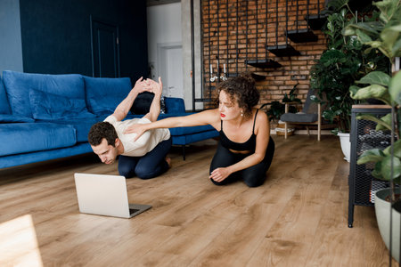Healthy couple stretching watching fitness video tutorial online on laptop. Young smiling fit man and woman doing yoga workout at home sitting on floor in living room together, hold handsの写真素材