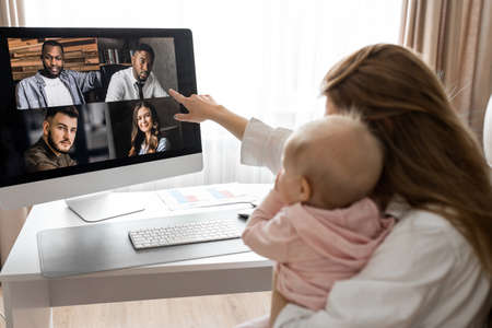 Lovely mother in maternity leave engaged in remote work or study at home on PC while infant baby girl hold documents and looking at screen. Modern day mother of little kid sit at desk use gadgets.の写真素材