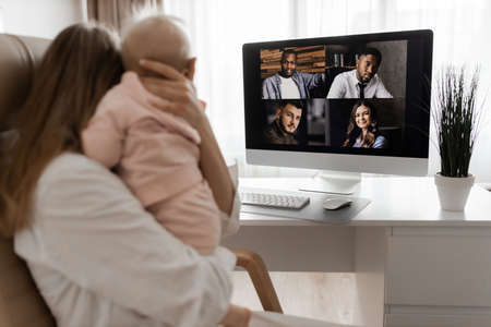 Blond young mother in maternity leave engaged in remote work or study at home on PC looking at...