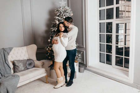Adorable marriage couple stand near Christmas tree at home, caring husband hug pregnant wife, young family excited about parenthood, enjoy happiness, winter holidays concept. high quality photoの写真素材