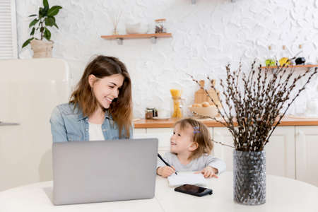 Beautiful single mother sitting at the desk with cute toddler, distantly working from home. Young business woman in maternity leave using laptop, searching in the internet ideas for new project.の写真素材
