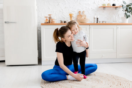 A strong and beautiful mother hugs her little daughter and goes in for sports in the morning in the kitchen. The concept of a healthy lifestyle. Training during quarantine with a small childの写真素材