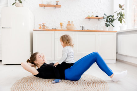 Beautiful smiling mother goes in for sports in the morning with her little daughter in the kitchen. Healthy lifestyle concept. Training during quarantine with a small child. High quality photoの写真素材