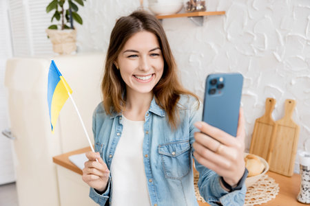 Young happy girl takes a selfie with the flag of Ukraine in her kitchen. Spot the war and peace in Ukraine, freedom for Ukraine. Yellow-Blue national flag of Ukraine. High quality photoの写真素材