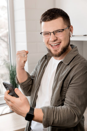 Happy young man looking at the camera, giving motivation to his co-workers while making a phone call, happy successful winner staying at home in the kitchen. The concept of working from homeの写真素材