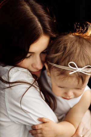 Dark-haired young mother gently holds in her arms and hugs little daughter on black background, happy caring mother embrace lull small baby girl child, enjoy tender moment, childcare concept.の写真素材