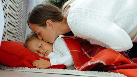 Young mother kissing her sleeping little daughter. Little cute girl sleeping cover plaid on the floor at xmas morning.の写真素材
