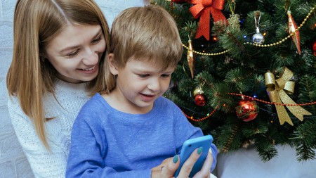 Young happy mother and son with mobile phone are sitting together near decorated christmas tree....