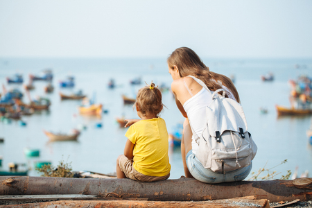 Young mother and her small daughter sitting on the seashore looking out over the ocean with fishing boatsの写真素材