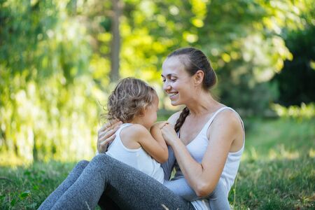 Mother and her little girl on green summer grass meadow having fun. Sporty mother and daughter having fun after yoga exercise outdoorの写真素材